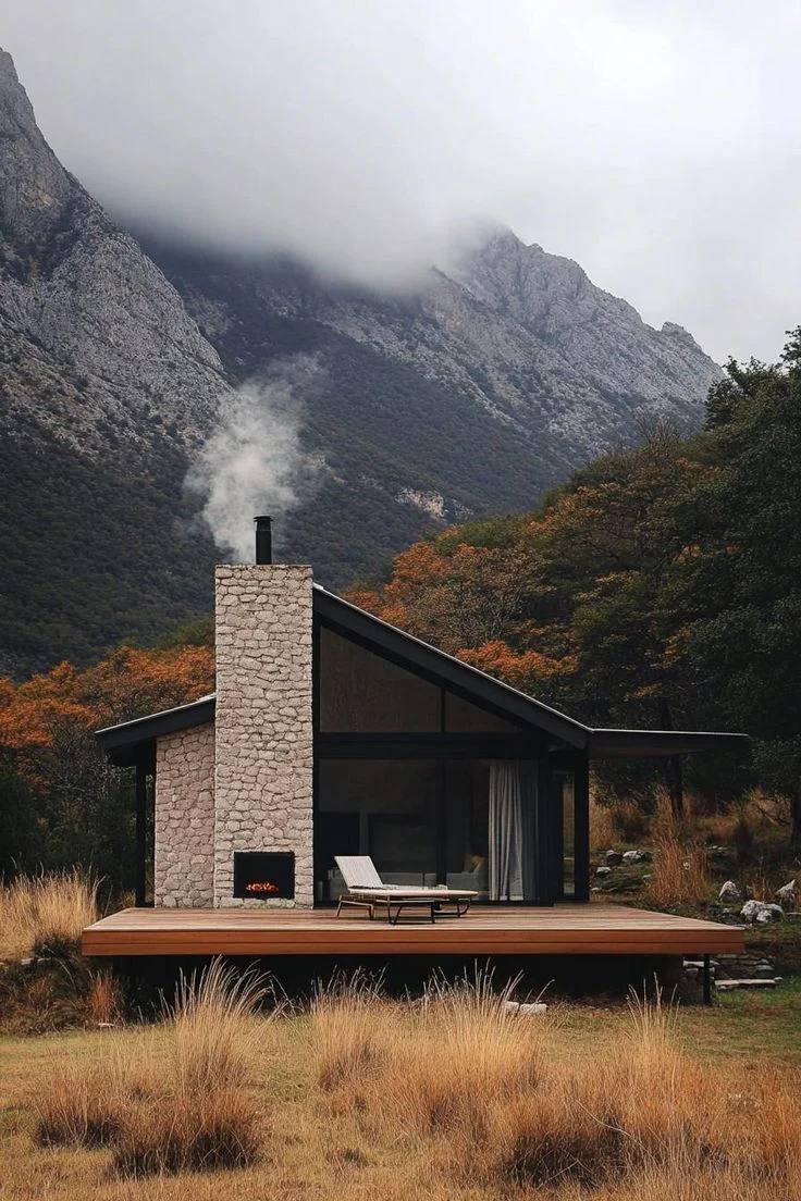 A-frame cabin with a stone chimney, smoke billowing from the top, a wooden deck with a lounge chair, and a view of mountains and trees in the background